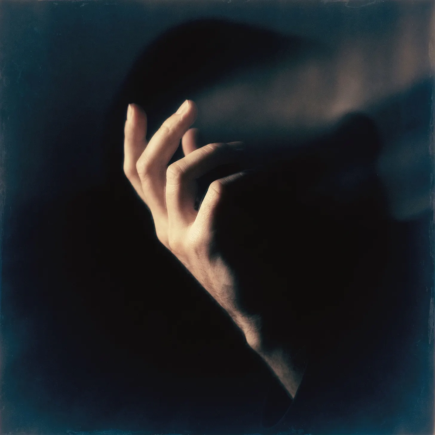 A softly lit, gently curled hand emerges from deep shadow against a dark, moody background, fingers relaxed and slightly parted, evoking a vintage, film-grain aesthetic.